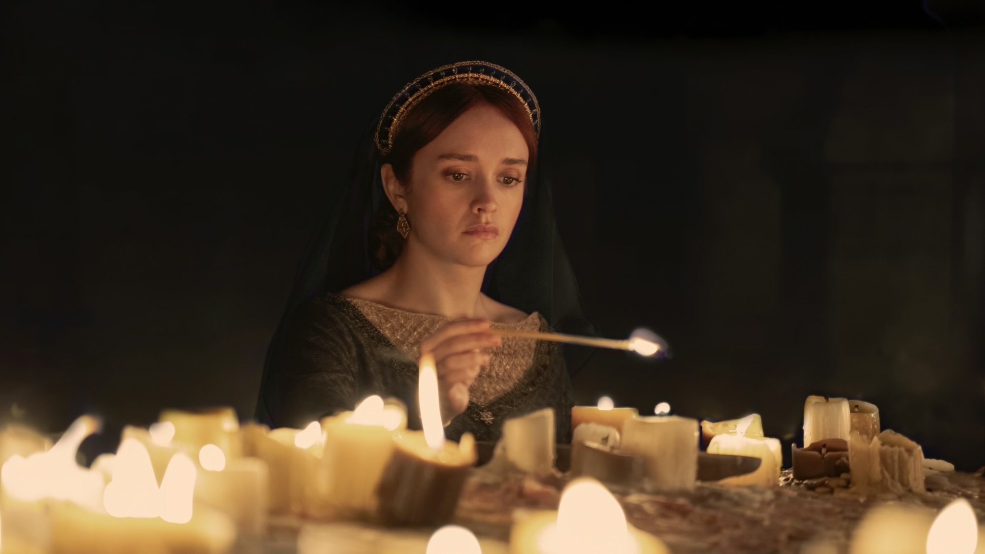 Alicent Hightower, wearing a green dress and black veiled headpiece, kneels at an altar and lights candles.