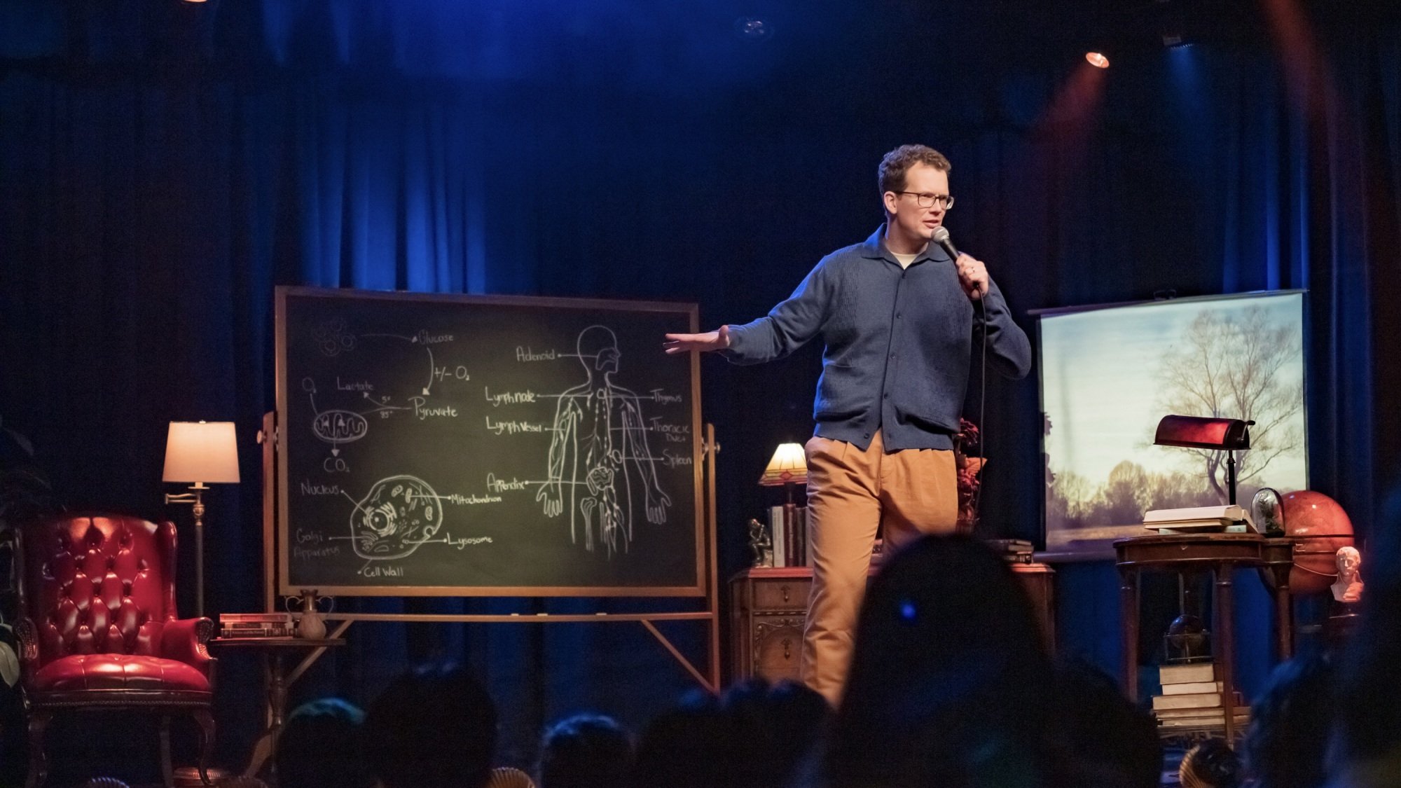 Hank Green performs stand-up onstage in front of a blackboard covered in diagrams of the human body and the cell.