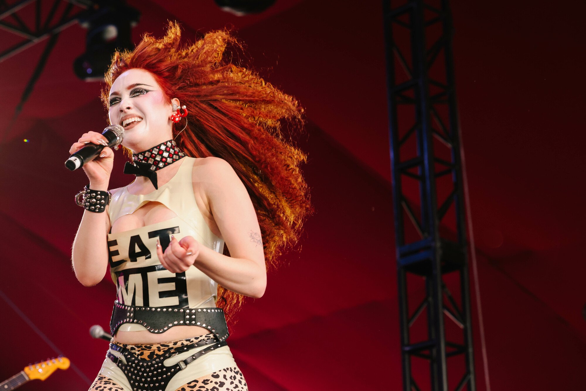 Chappell Roan, with wild red curly hair, wearing an "EAT ME" shirt and holding a mic on stage at Coachella.