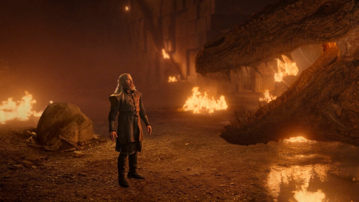 A bearded man stands in front of a dragon in a burning cave.