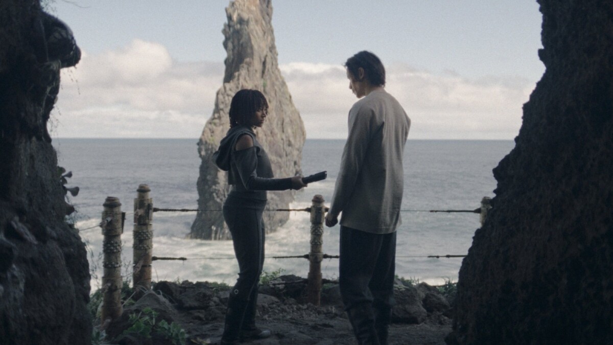 Osha holds an unlit lightsaber between her and the Stranger; the two stand in a cave mouth overlooking the ocean.