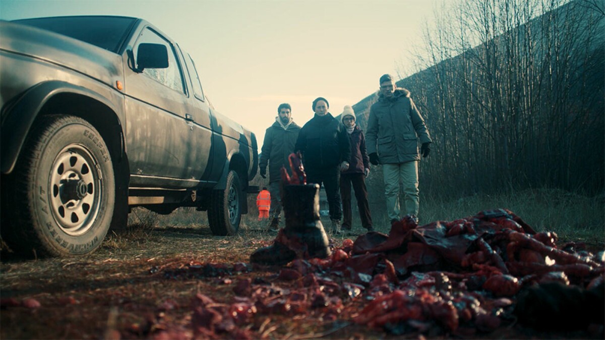 A group of people next to a car stand staring at a pile of human remains with a shoe standing next to it.