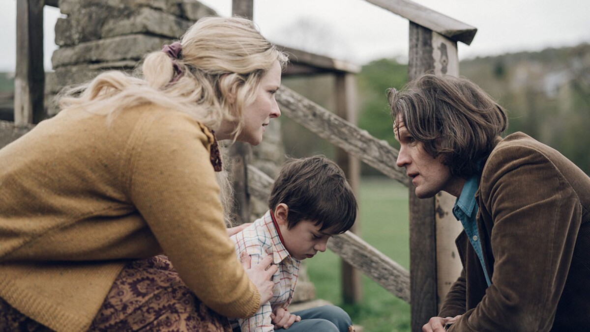 A woman, a man and a little boy crouch in the countryside beside a fence.