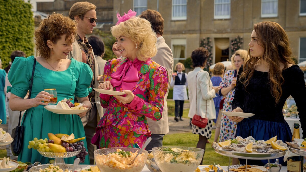 Three women in '80s costumes stand beside a table laden with food at a party in the TV show "Rivals".