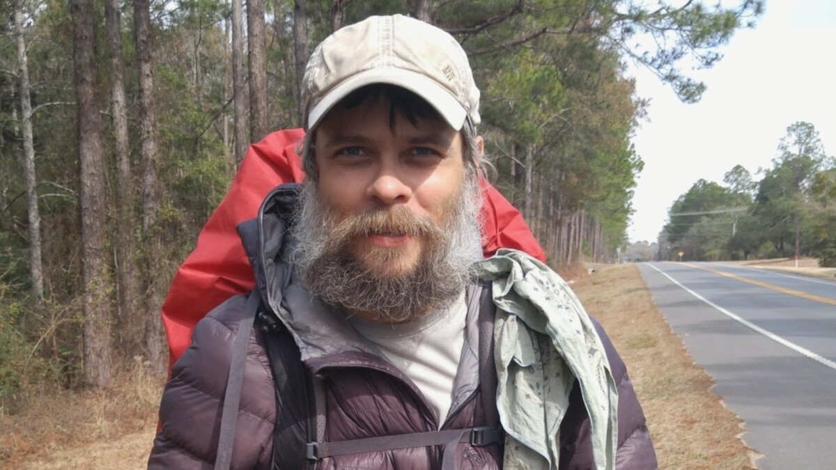 A hiker with a beard stands by a roadside. 