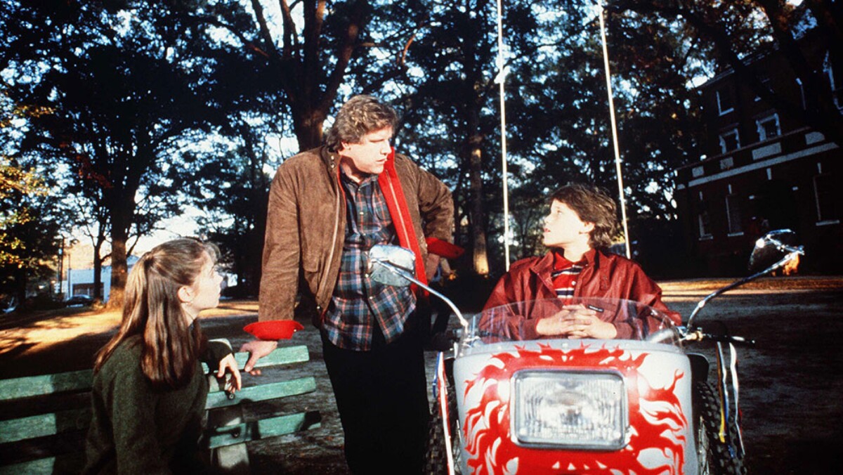 A girl sits on a bench beside a boy standing and another boy sitting on a wheeled vehicle.