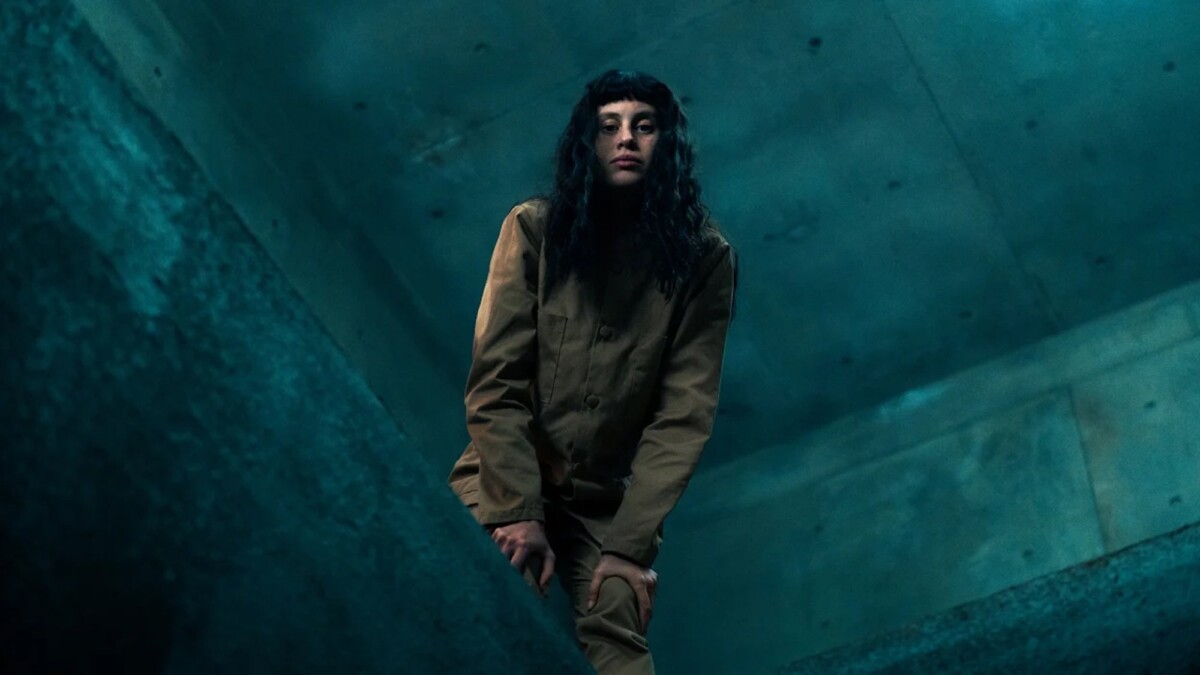 A woman in a prison uniform leans over a platform edge.