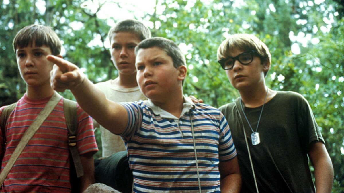 Four boys stand in the woods with bags over their shoulders, one points at something for the others to look at.