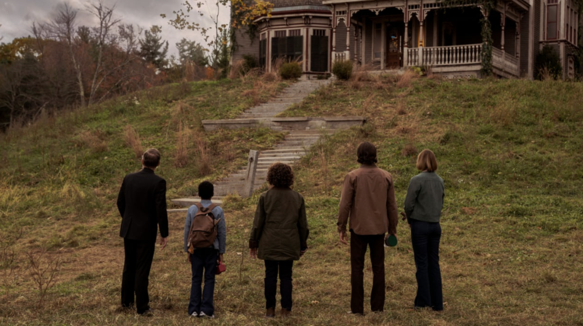 A group of people stand at the foot of a hill, looking up at a large, creepy house.