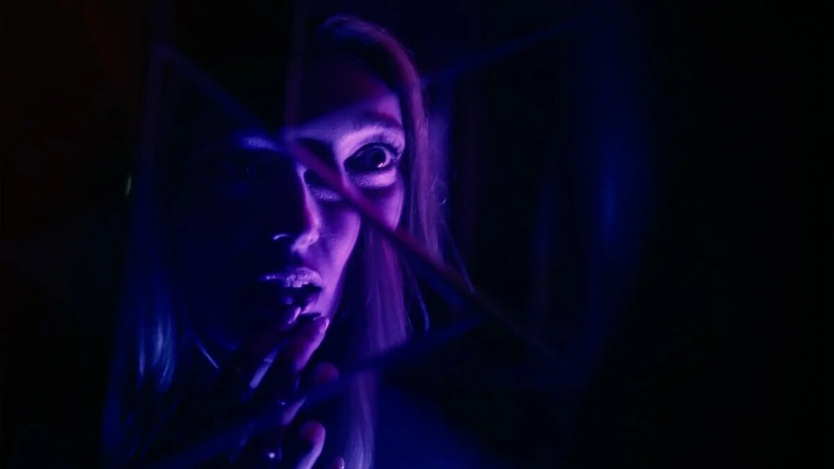 A purple-lit image of a woman looking into a smashed mirror.