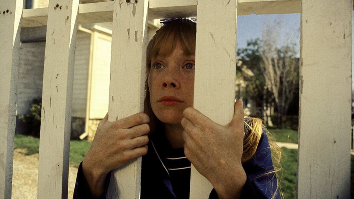 A sad looking girl peers through wooden slats.