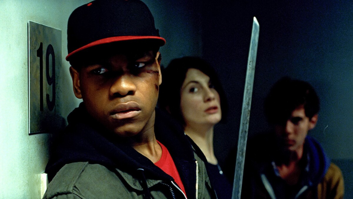 Three people stand in a corridor, looking scared. The man closest to the camera holds a sword and his face is cut.