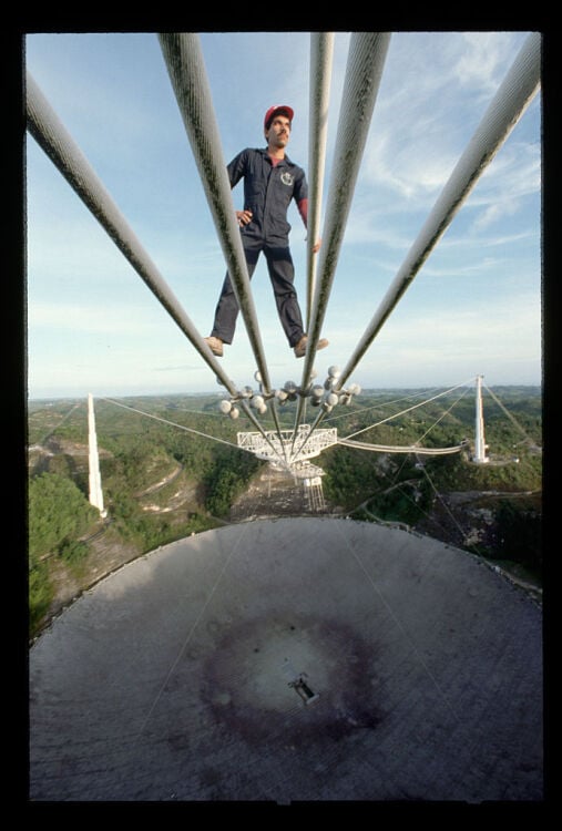 An archival photo shows a technician standing atop cables over the Arecibo telescope about 450 feet in the air.