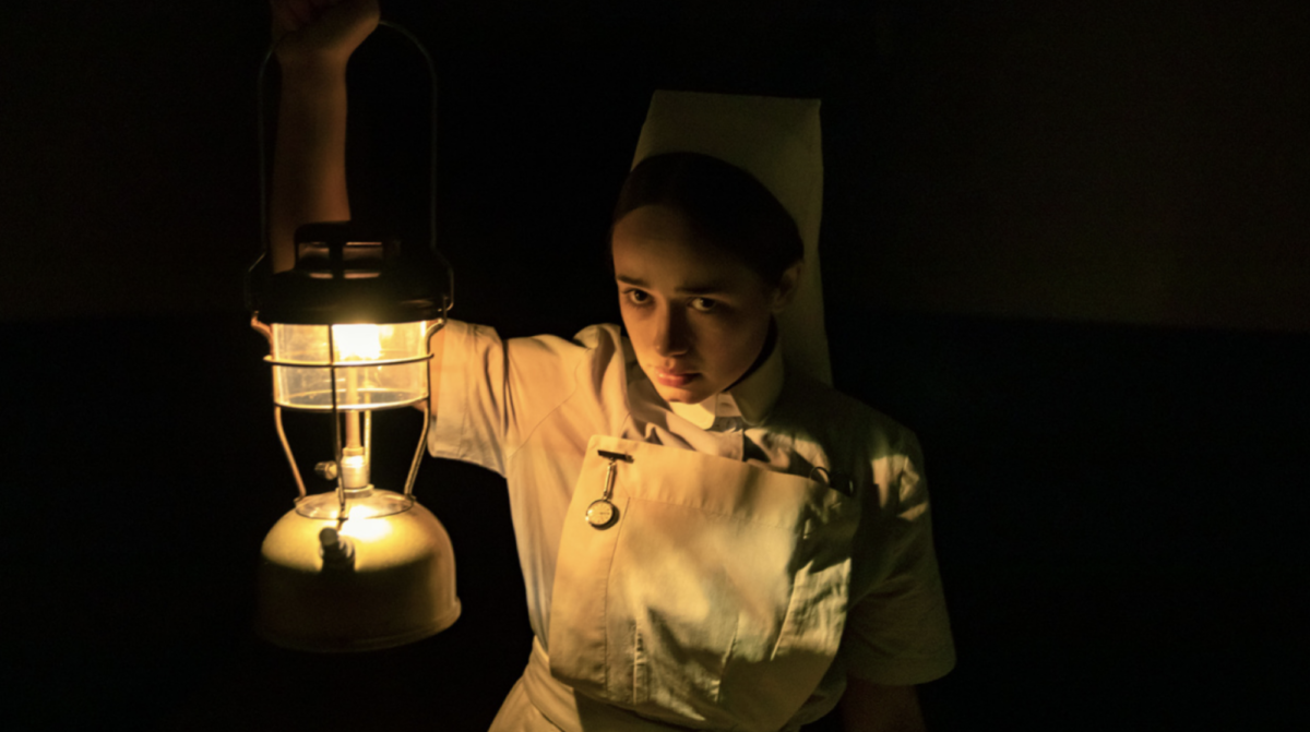 A woman in an old-fashion nurse's uniform holds up a lantern in a dark room.