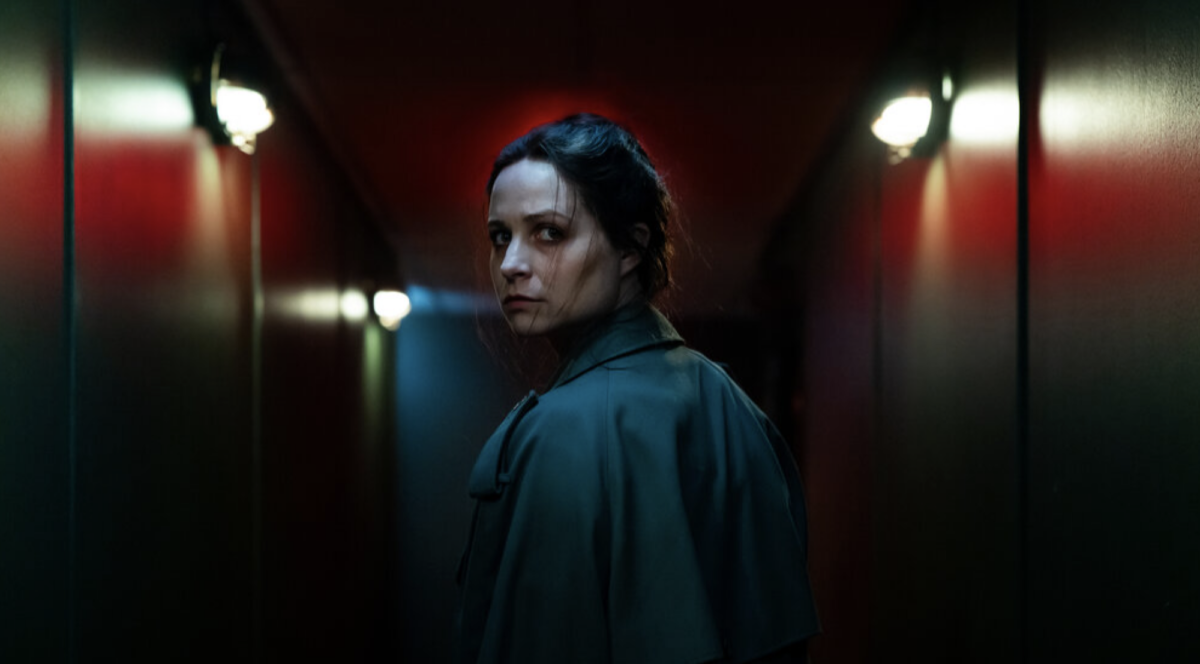 A woman walking down a dark corridor looks back over her shoulder.