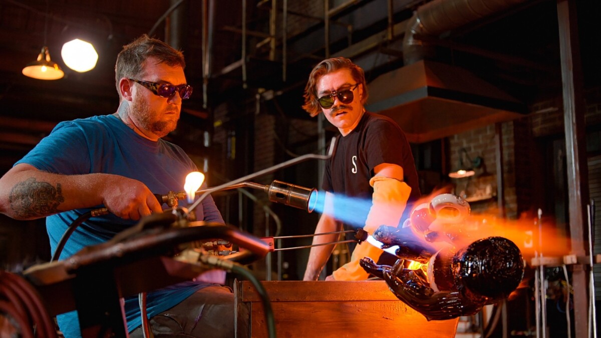 Two men wearing protective goggles blow glass.