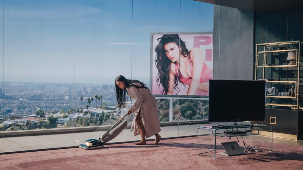 Demi Moore vaccuums while a billboard featuring Margaret Qualley looks on in "The Substance."